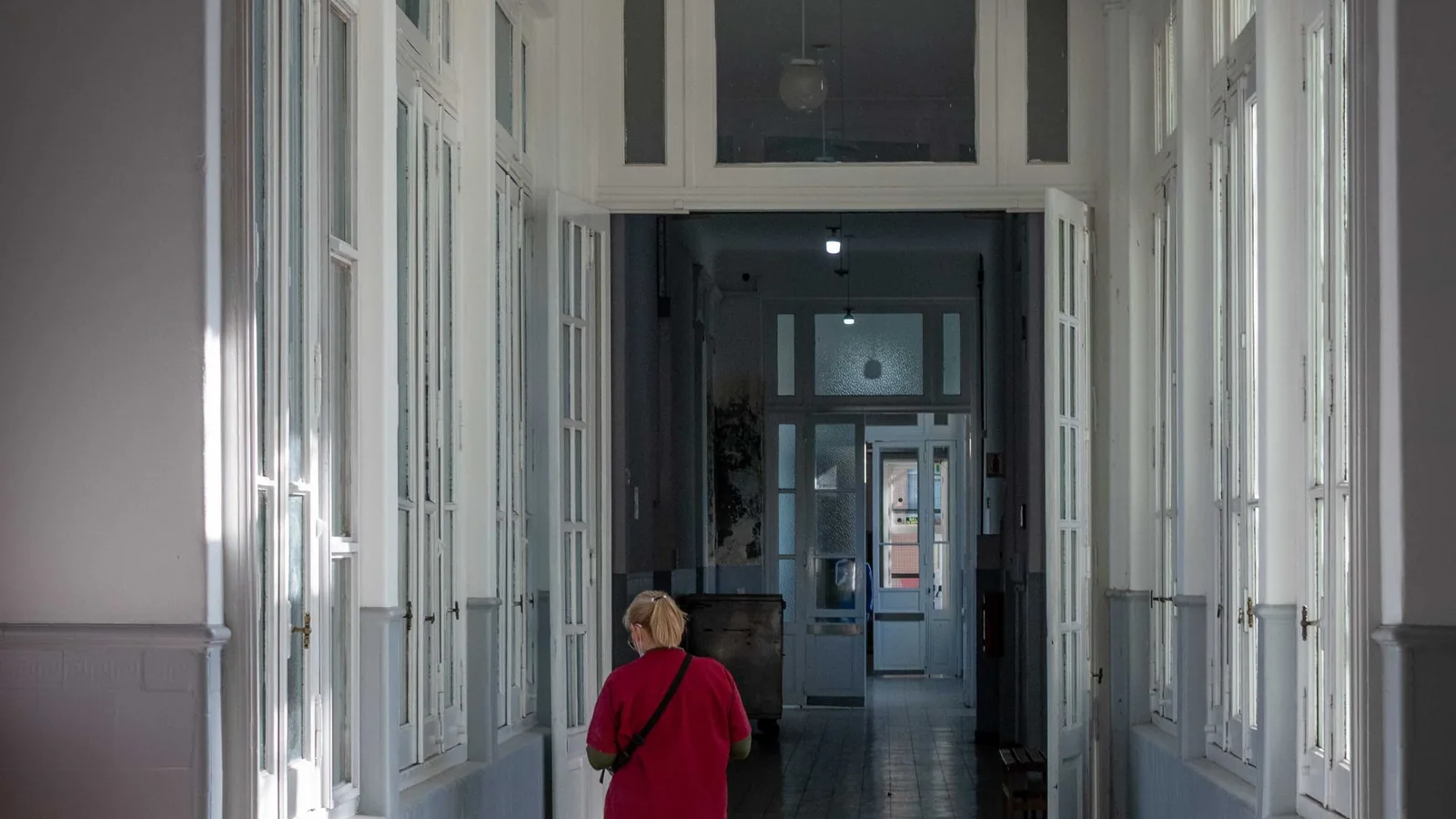 A nurse in red scrubs stands in a hospital corridor looking at a phone, with a glass door and a wall sign behind her.