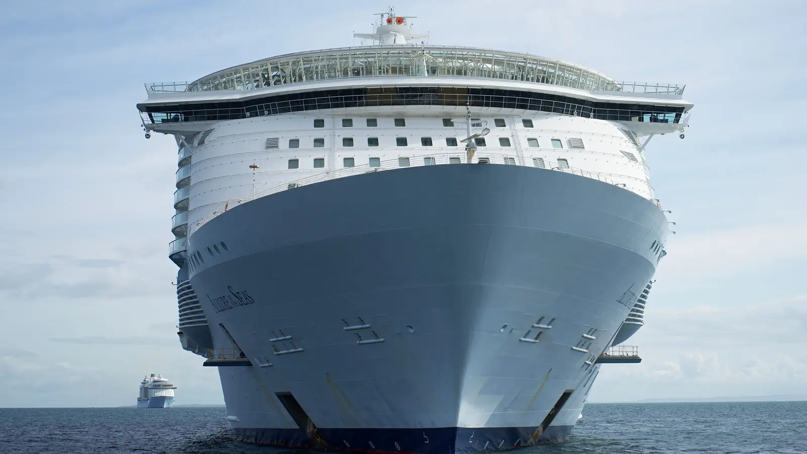 A large white cruise ship seen from the bow, with stacked decks, orange lifeboats, and a glass upper structure, moving across deep blue water under a pale sky.
