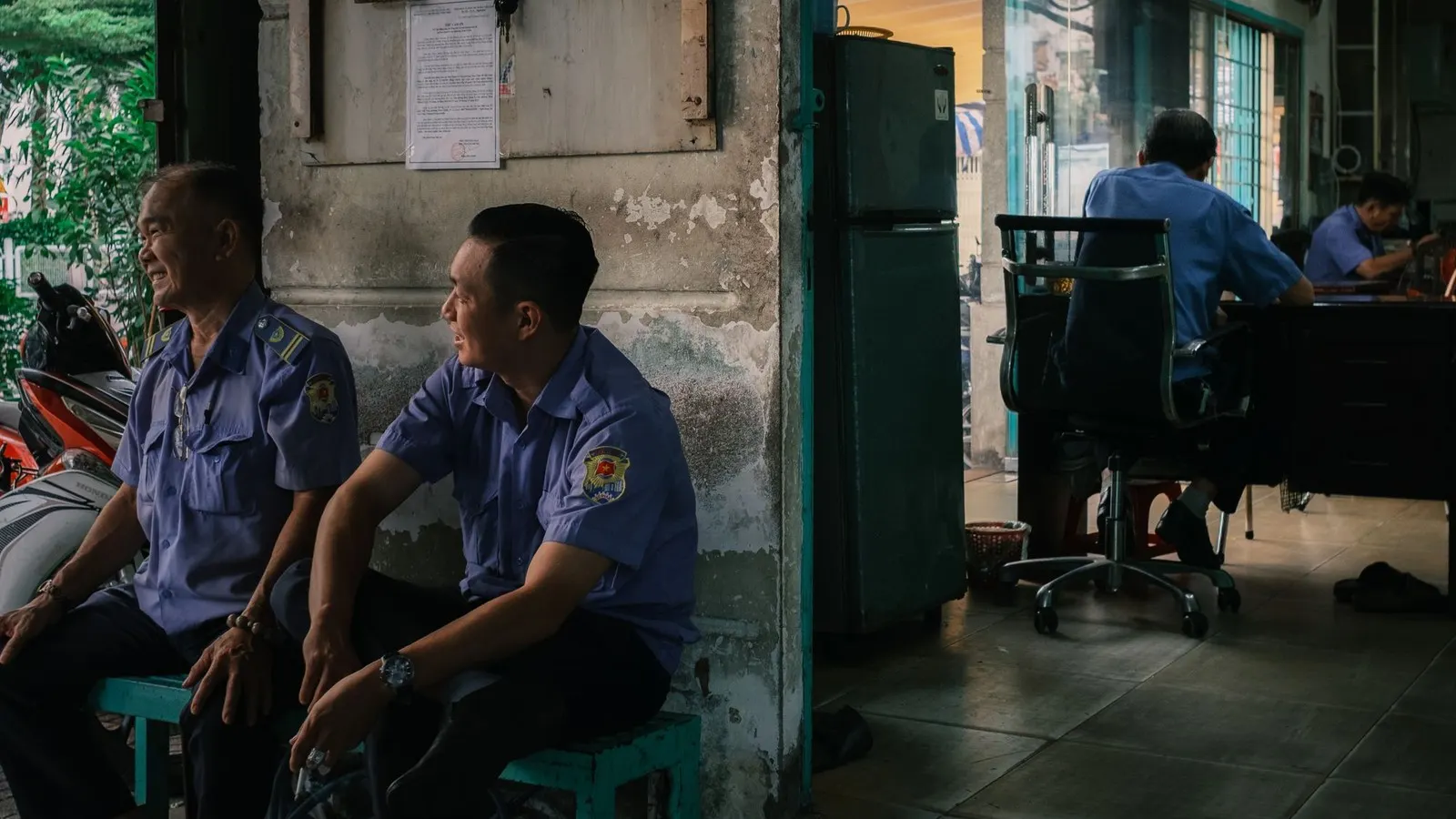 Three people in dark blue uniforms in a security office: one sits at a laptop on a wooden desk, and two stand nearby looking at phones, with a window and a wall of small photographs behind them.