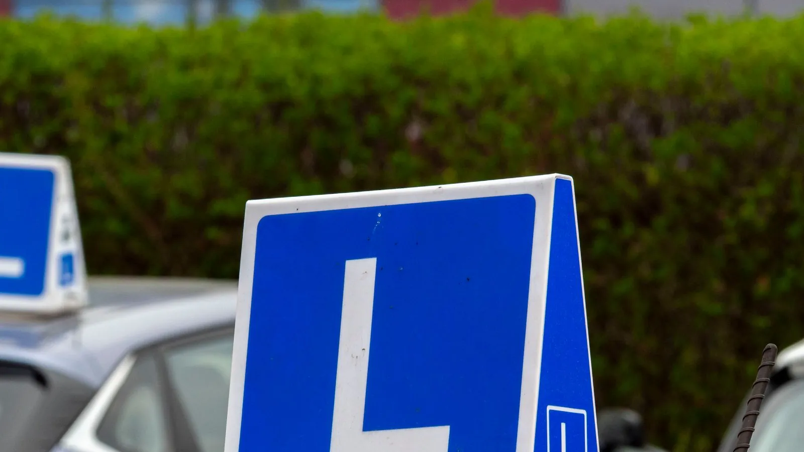 A white hatchback and a white SUV parked on gravel at a driving school, each with a red learner plate on the front bumper.