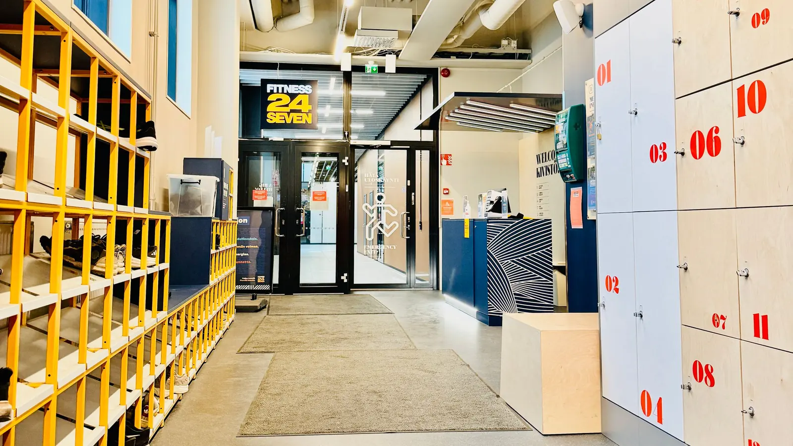 A person in black athletic wear walks across a black mat toward a light-wood reception desk in a gym entrance, with wooden lockers on the left and a glass wall behind the desk.