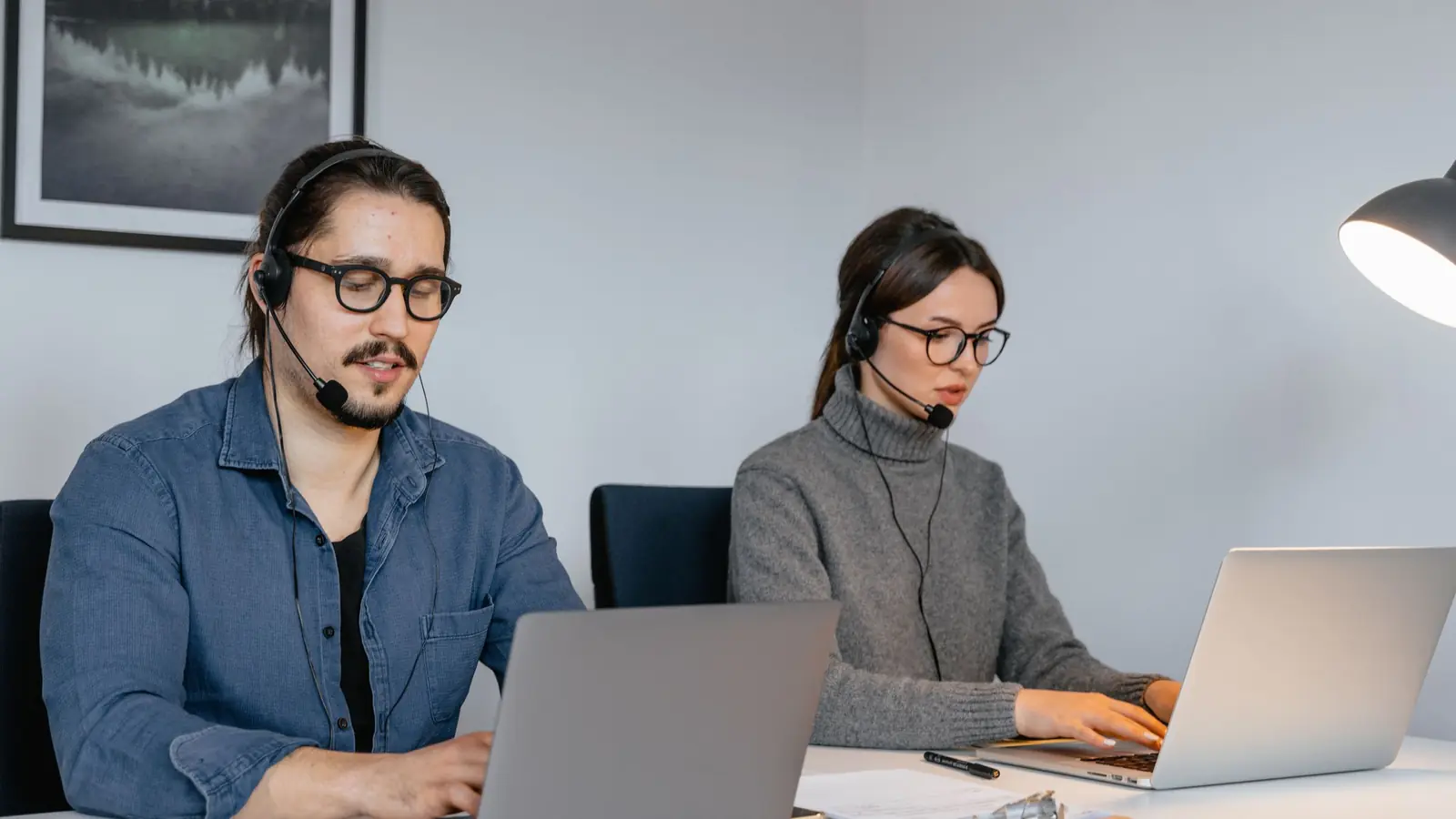A man and a woman wearing headsets sit at laptops in an open office, with a glass partition and more desks behind them.