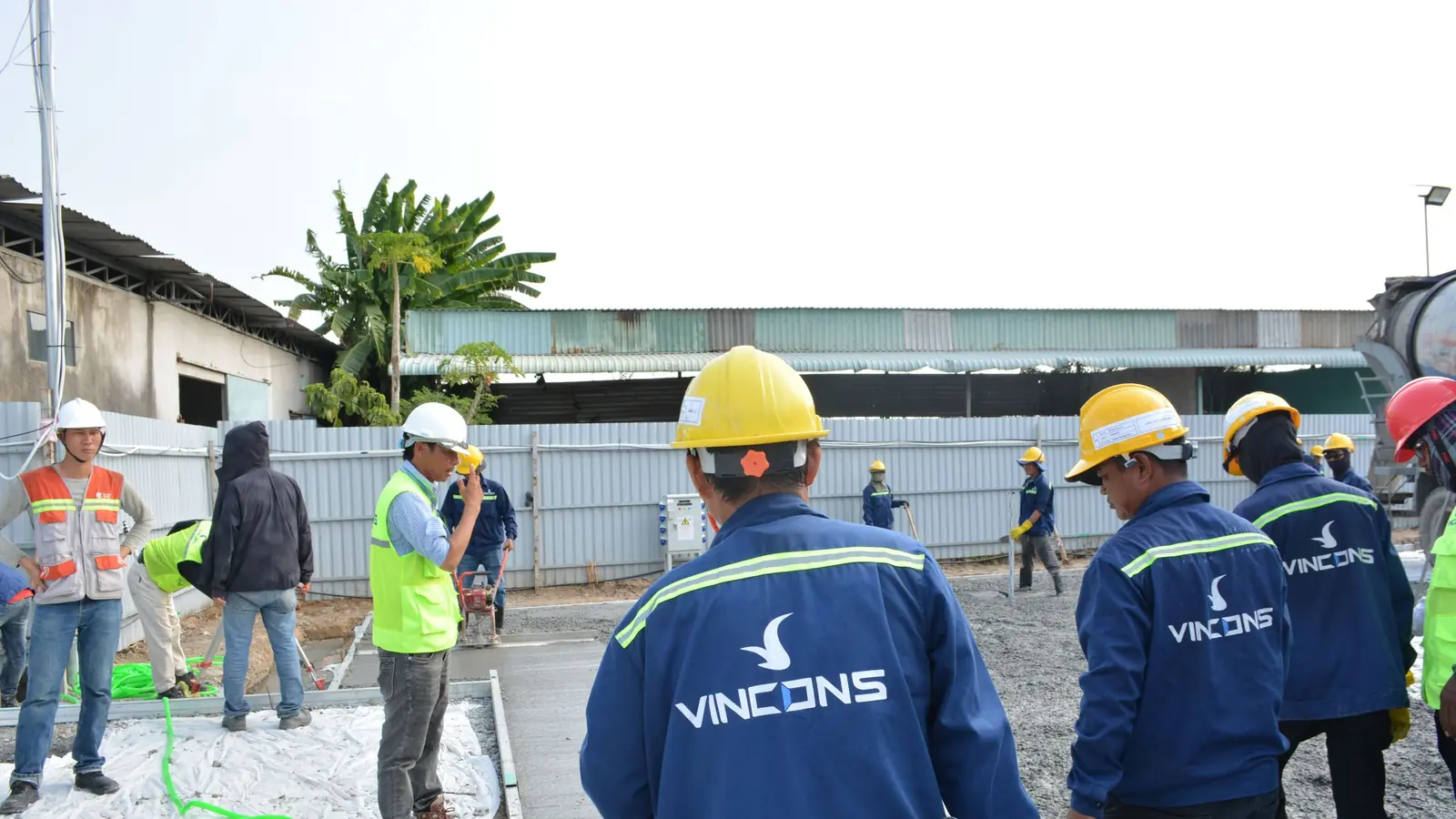 Five construction workers in yellow hard hats and orange high-visibility vests stand together on gravel, with a concrete building frame and an excavator behind them.